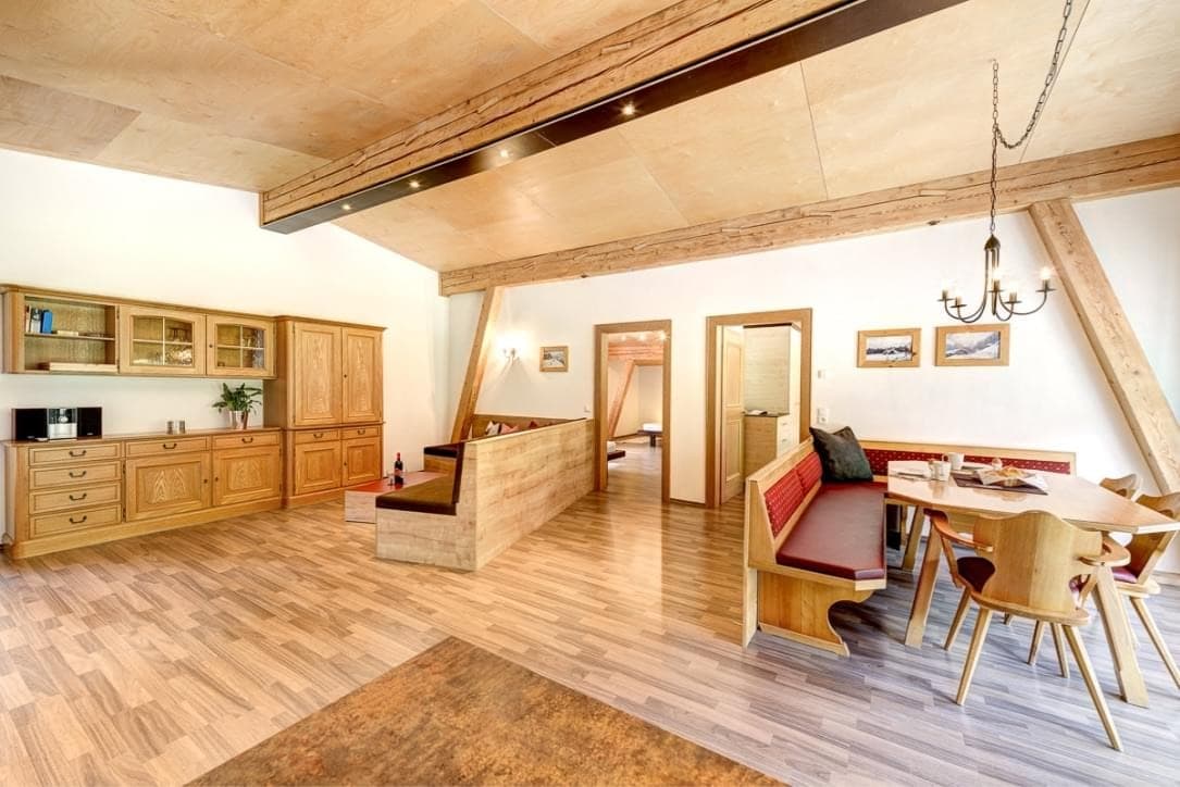 Bocks Apartments 61 - Top 2 Ursprung - A bright bedroom with a large bed, wooden beams on the ceiling, and wooden floors. Large windows offer a view of green trees outside. A red sofa is in the foreground.